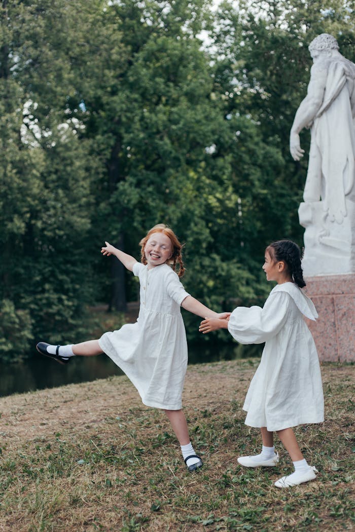 Two children dance with joy in a park, wearing white dresses beside a sculpture.