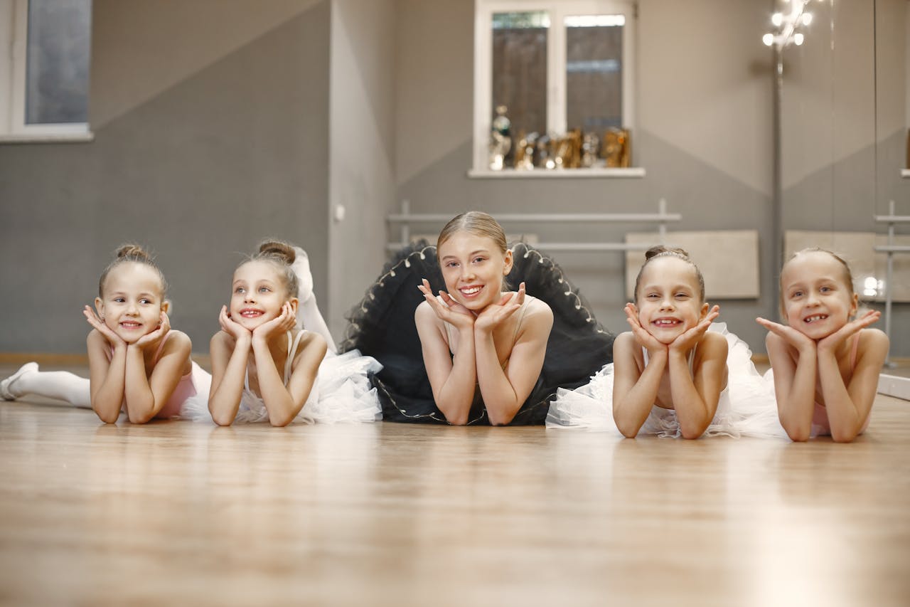 services-04 Five young ballerinas lying on the studio floor, smiling at the camera in tutus.